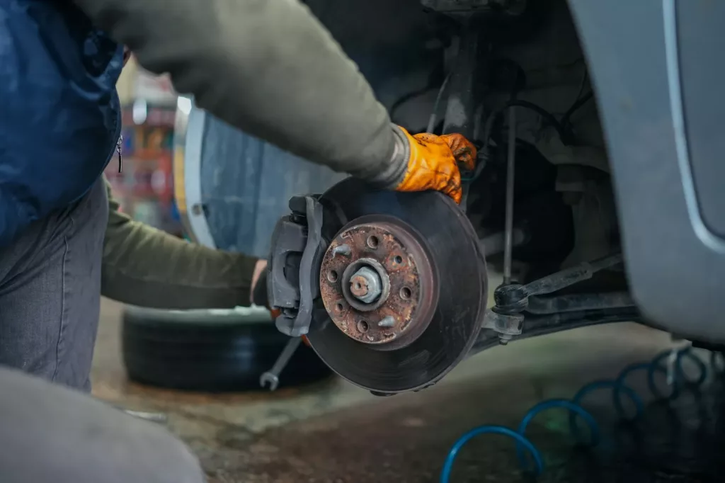 Mechanic in an orange work glove reaching toward an exposed brake rotor and caliper with the wheel removed and a tire on the shop floor — brake repair and inspection at Rite-Price Auto Service Centre in Calgary, AB