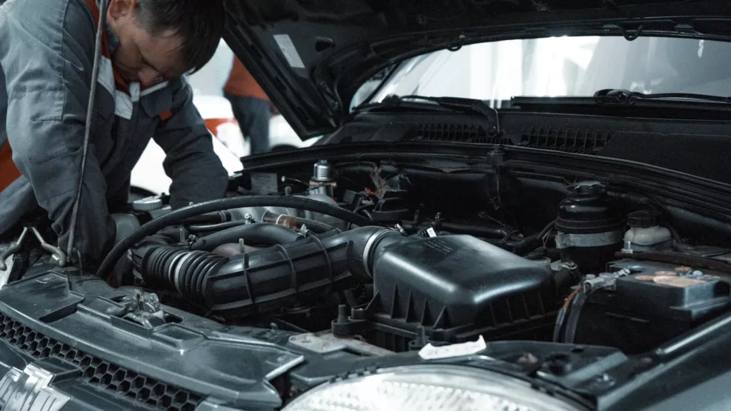 Mechanic in grey coveralls leaning under the open hood of a vehicle inspecting the engine bay — routine car maintenance and service at Rite-Price Auto Service Centre in Calgary, AB