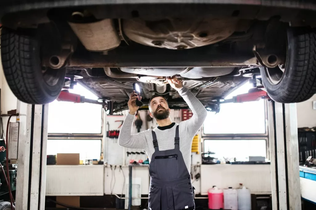 Mechanic inspecting the underside of a vehicle on a hoist with a work light — part of an Alberta out-of-province vehicle inspection at a Calgary auto shop, Rite-Price Auto Service Centre